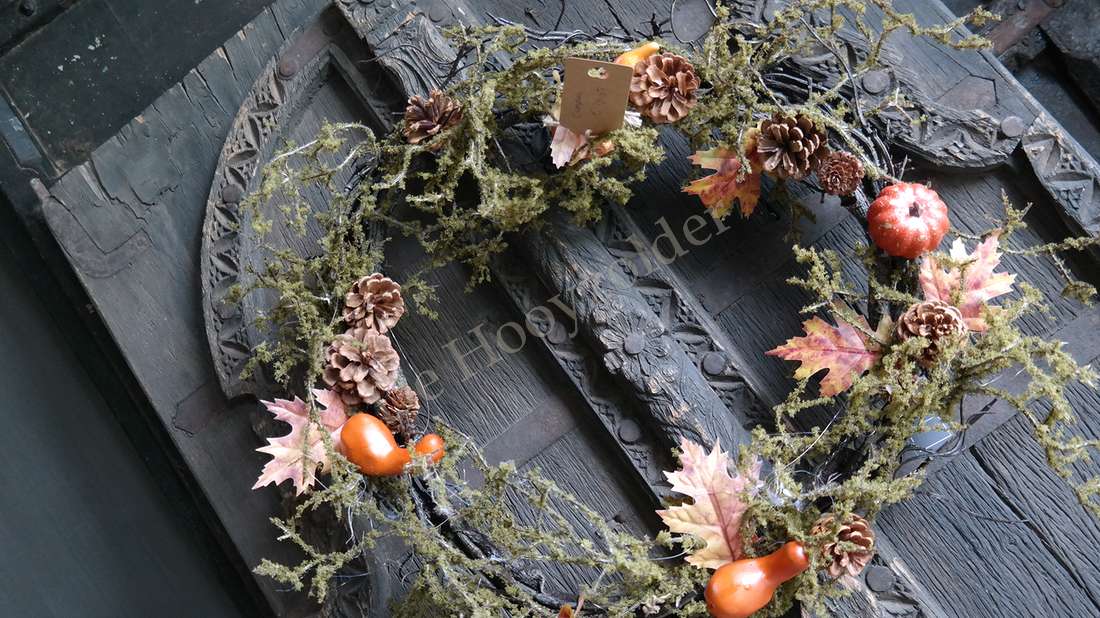 Mos-herfstkrans van De Hooyzolder met dennenappels, herfstbladeren en oranje pompoenvormige decoraties aan een donker houten deur