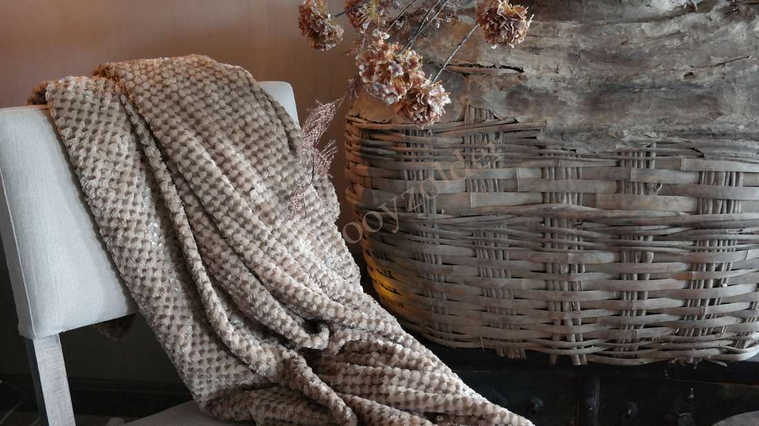 Plaid Rick fluffy - nougat, pluizige beige deken drapeert over een witte stoel naast een rieten mand