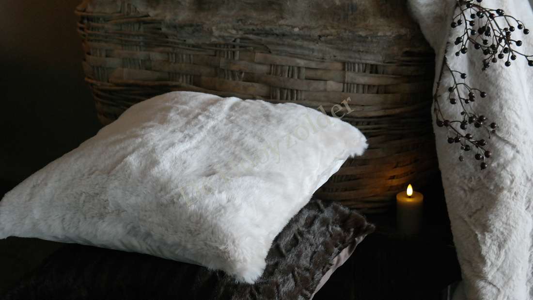 Kussen Zaya bont créme, wit-creme faux fur kussen op wicker achtergrond bij een brandende kaars