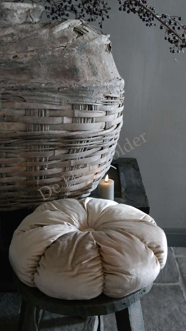 Kussen velvet rond beige van De Hooyzolder; ligt op een houten kruk naast een grote rieten mand.