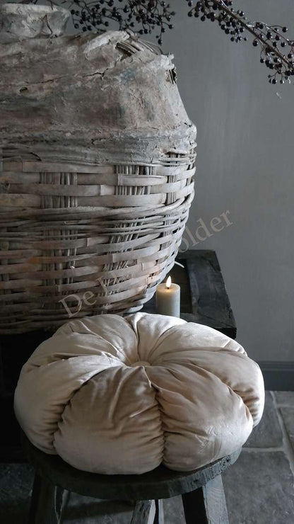 Kussen velvet rond beige van De Hooyzolder; ligt op een houten kruk naast een grote rieten mand.