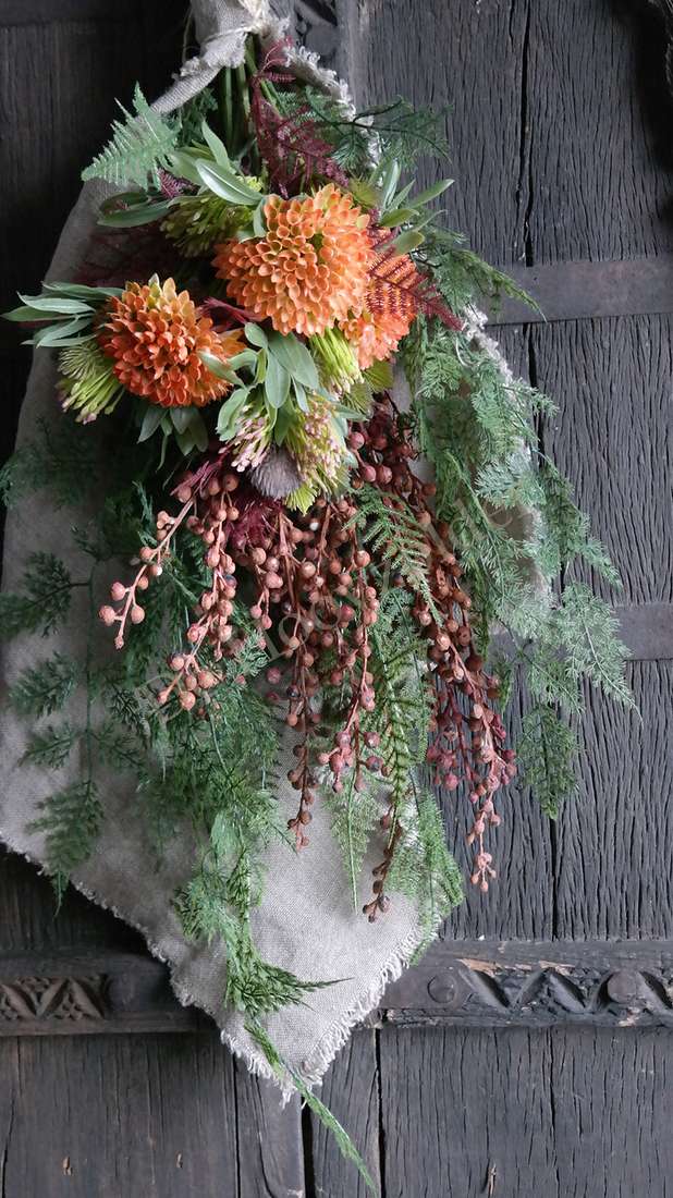 Kunsttak dadeltak oranje met groen blad en bruine hangende trosjes, bevestigd aan een beige doek tegen een houten muur.