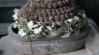 Ovaal houten olijfbak met grijze patina en touwrand, gevuld met gedroogde bruinachtige zaaddozen en groene bladeren.