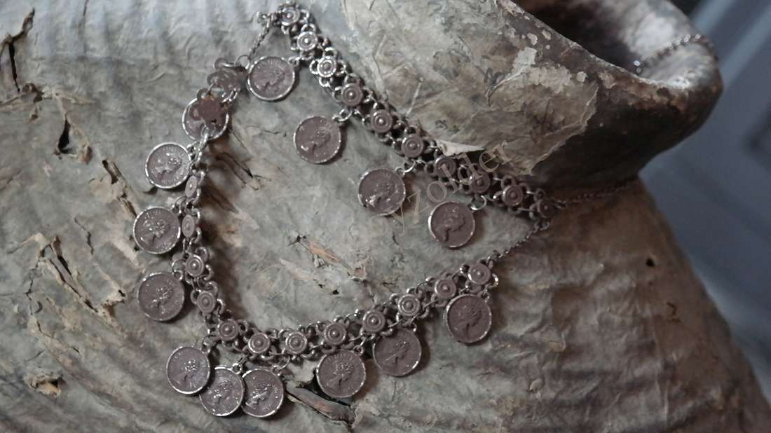 Decoratieve sierketting met zilveren muntjes aan een dubbele ketting, liggend op een ruwe achtergrond.