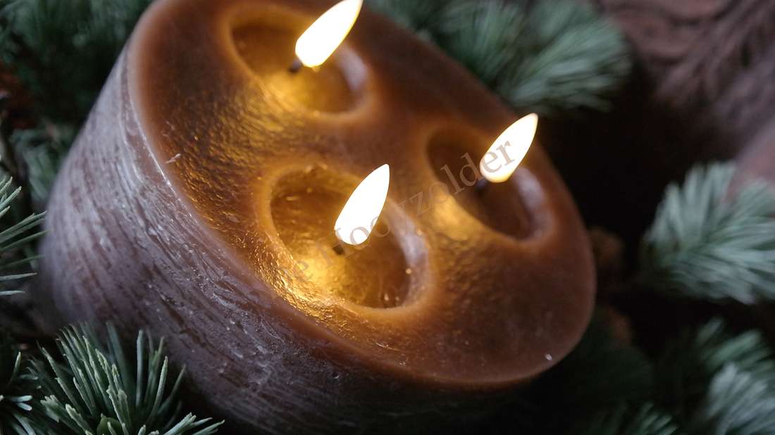 Donkerbruine LED-stompkaars met drie lonten in ronde keramische houder, brandend tussen dennen takken.