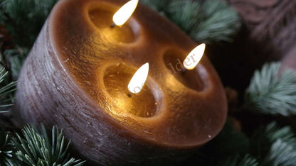 Donkerbruine LED-stompkaars met drie lonten in ronde keramische houder, brandend tussen dennen takken.
